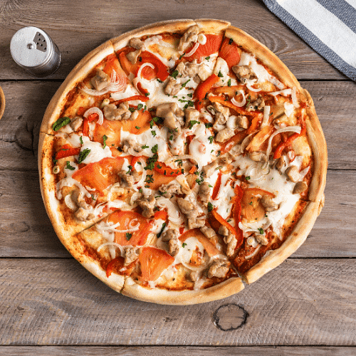 Pizza with chicken, tomatoes, onions, and herbs on a wooden table with a napkin and pepper shaker.