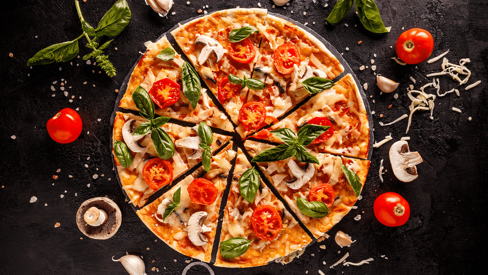 Pizza topped with tomatoes, basil, mushrooms, and cheese on a dark background with ingredients scattered.