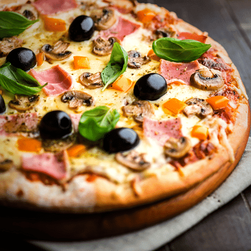 Close-up of a freshly baked pizza with olives, mushrooms, ham, bell peppers, and basil leaves.