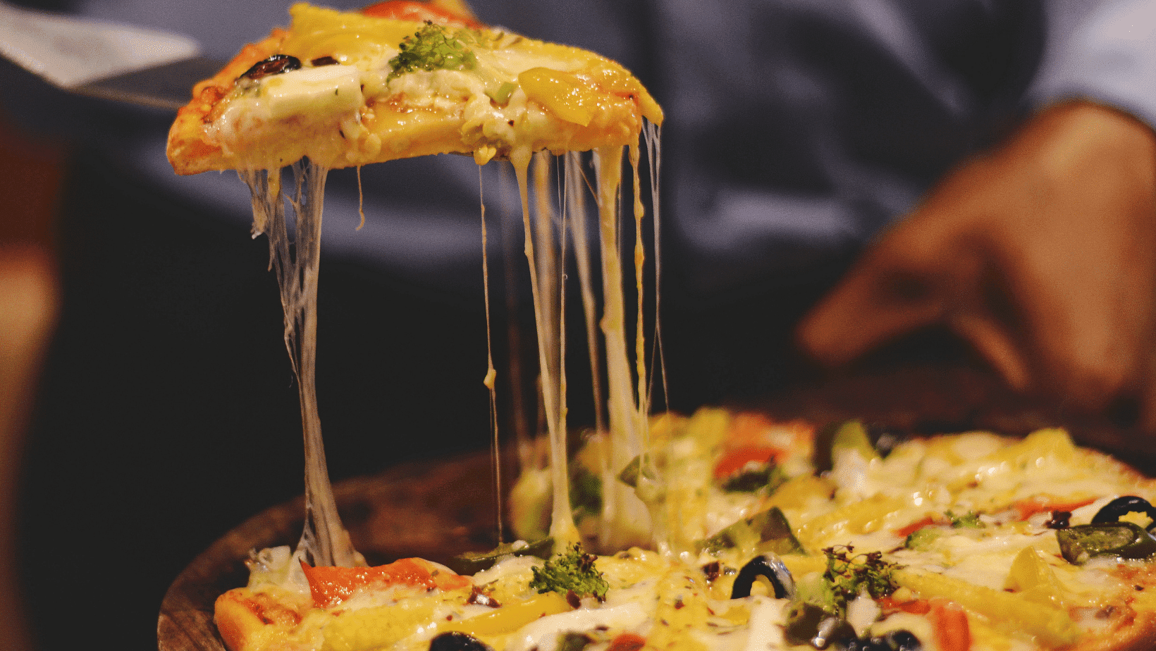 A slice of cheesy vegetable pizza being lifted from the pan, showing melted cheese and toppings.